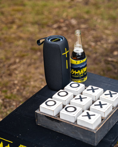 camping table holding bluetooth speaker, drink, and game of naughts and crosses